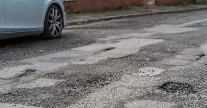 A South Cheshire road surface broken up by several potholes, with a silver car parked at the kerb beside a brick wall.